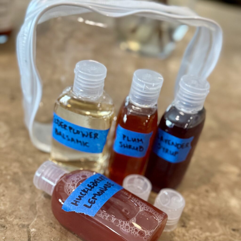 Four small clear flip-cap travel bottles filled with colorful liquids and labeled with blue tape, sitting on a counter with a clear zippered pouch in the background.