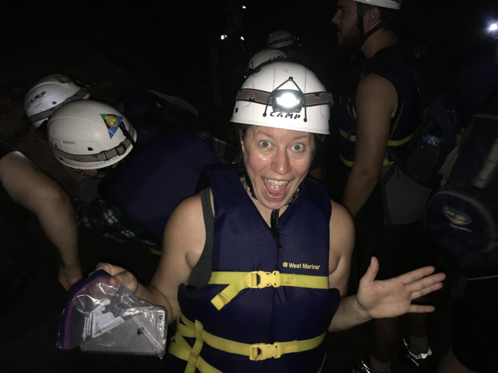 Ginny Dee wearing a helmet and life vest, holding a phone sealed in a plastic bag with an exaggerated grin — travel health and safety with a cheeky twist.