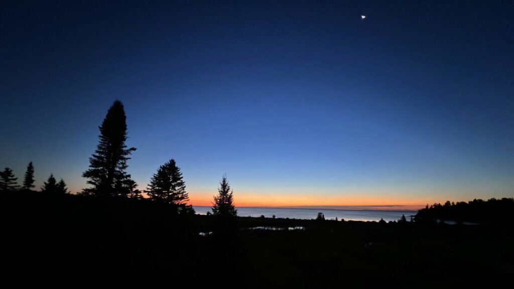 Silhouetted pine trees at dawn with a crescent moon and glowing horizon over a calm lake — a peaceful, expansive view of sky and water.