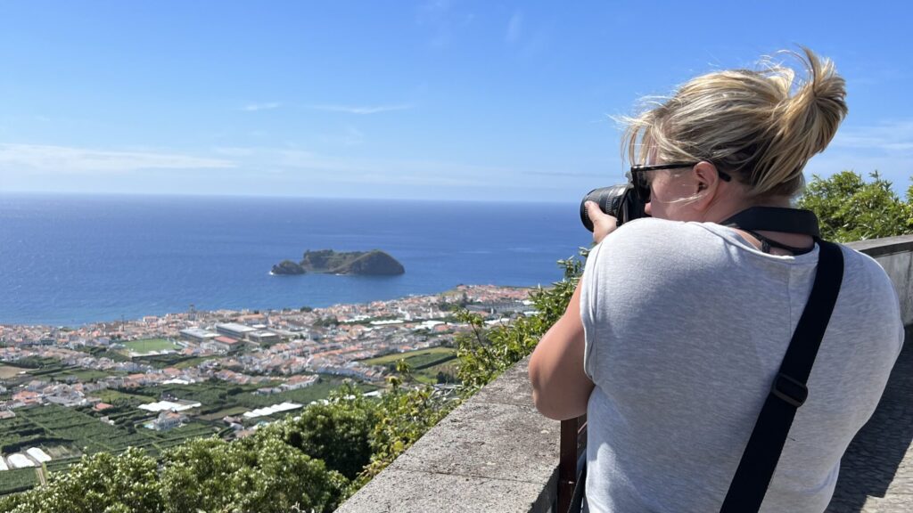 Ginny stands on a church balcony in the Azores with her back to the camera, taking a picture with her DSLR.  In the background we can see a village overlooking the sea.