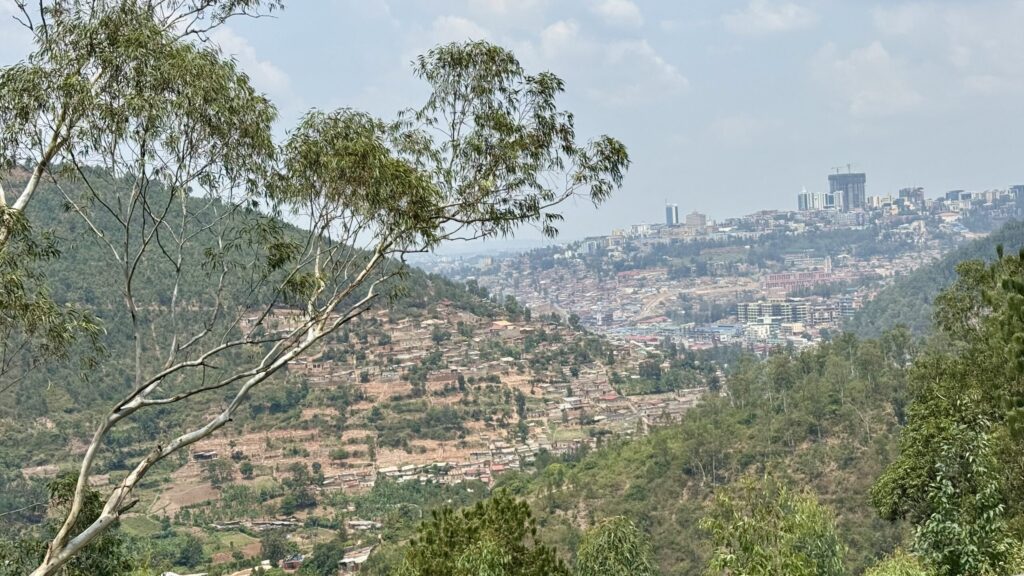 A vista view of Kigali in the distance, skyscrapers topping the hill, fronted by two crossing hills covered with forrest and the modest, tan buildings of an older town.