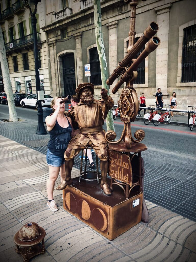 Ginny looks through a telescope on Las Rambles with a street performer dressed as Galileo's Statue.