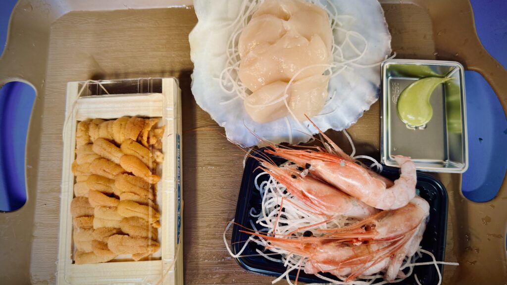 A tray of fresh, raw seafood from Tsukiji Market in Tokyo - a large tray of Uni, thinly sliced scallops served in their shell, and sweet shrimp, along with a dollop of wasabi paste.