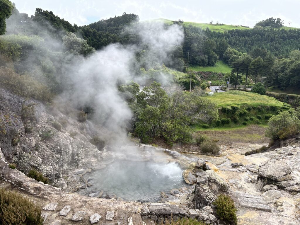 Steam rises out of a volcanic vent in the town of Furnas on Sao Miguel Island in the Azores, in front of rolling green hills covered in thick trees.