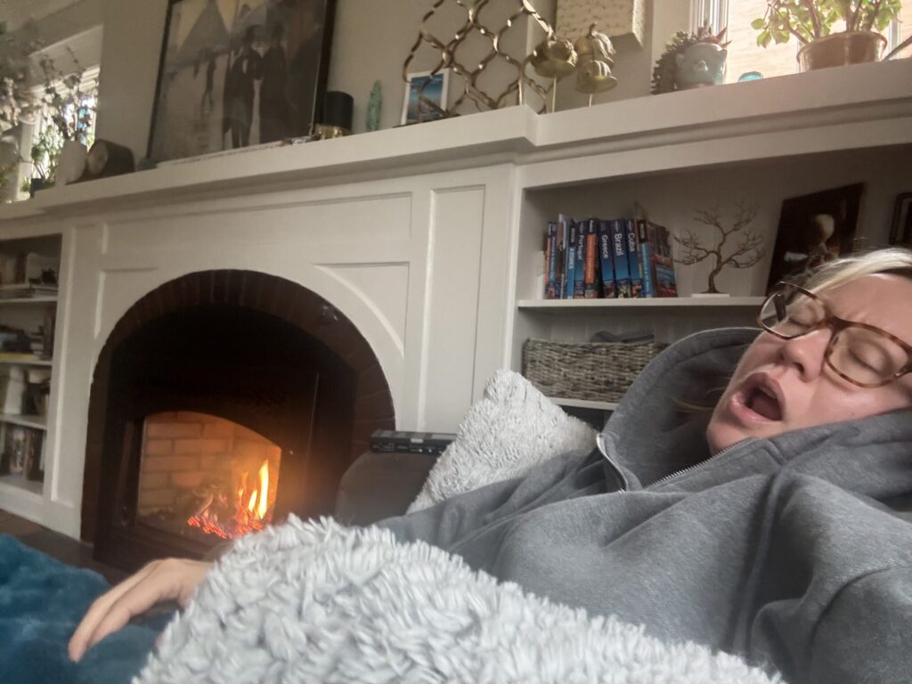 Ginny napping by a lit fireplace, wrapped in blankets and a hoodie on a couch.