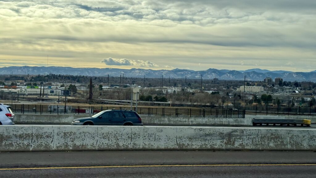 Cars driving along a snowy winter highway with mountains in the distance under a dramatic gray sky.
