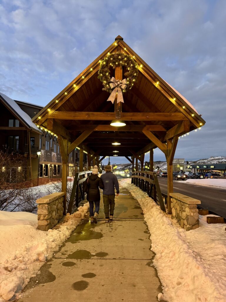 Couple walking under a wooden pedestrian bridge decorated with a holiday wreath and warm lights.