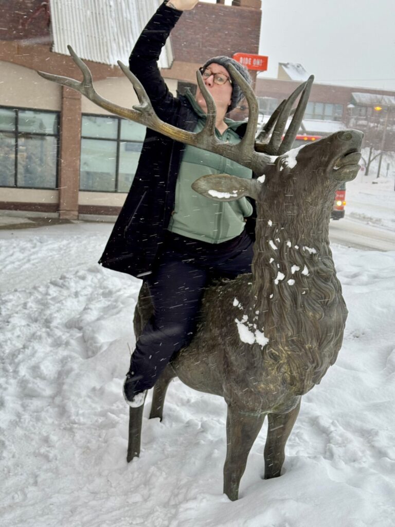 Ginny posing dramatically while riding a snow-covered elk statue during a winter storm.