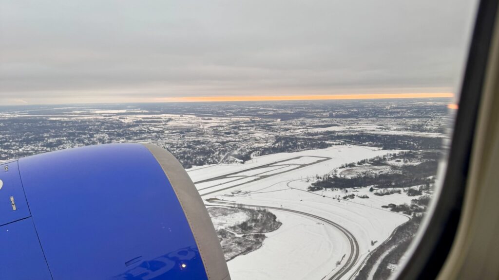 Aerial view from a plane window of snowy fields and winding roads beneath a gray sky.