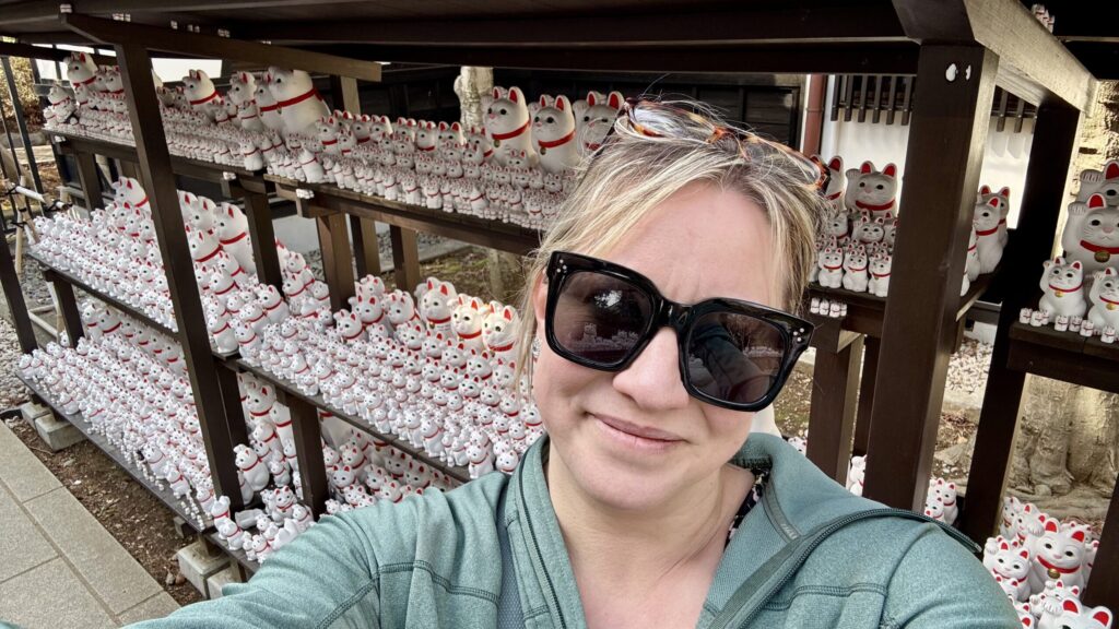 Ginny smiling in front of approximately 5,297 waving cat statues of all sizes, at Maneki-Neko Shrine in Tokyo.